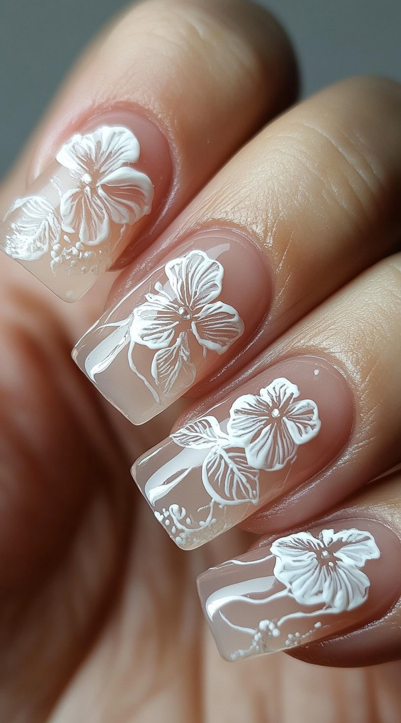 A hand with square-shaped nails featuring a sheer nude base adorned with intricate 3D white floral designs.