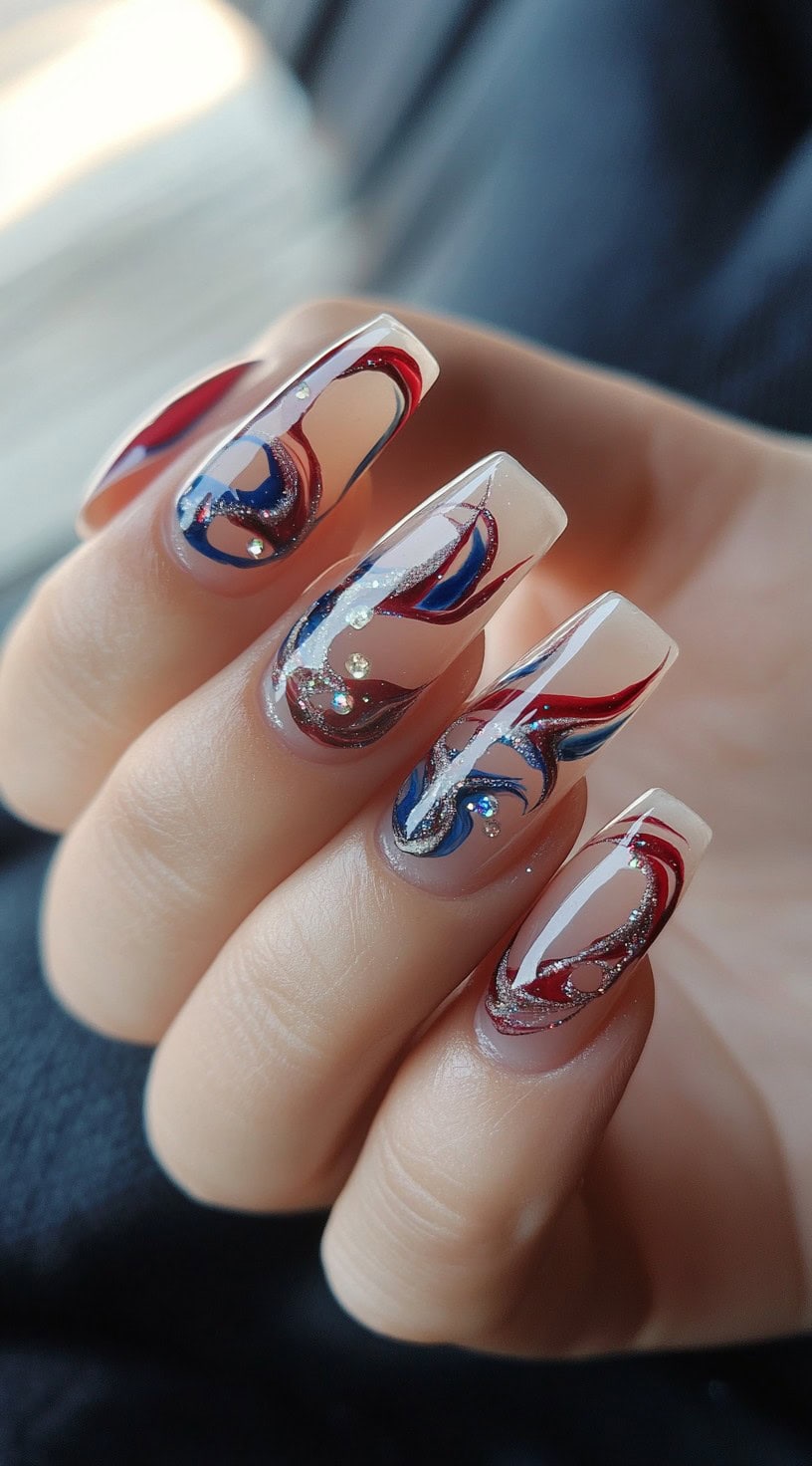A hand featuring long coffin-shaped nails with an abstract red and blue swirl design over a sheer nude base, accented with fine silver glitter and rhinestones.