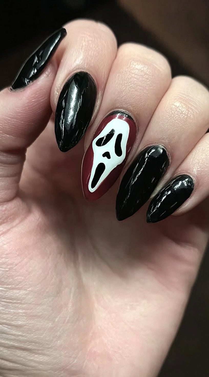 Hand with almond-shaped nails painted black, featuring one nail with a burgundy base and a white ghostface design.