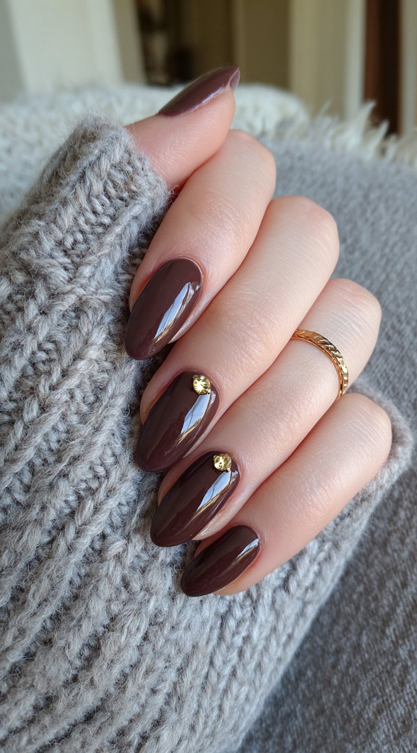 Almond-shaped nails painted in glossy chocolate brown with small gold rhinestones on two nails.