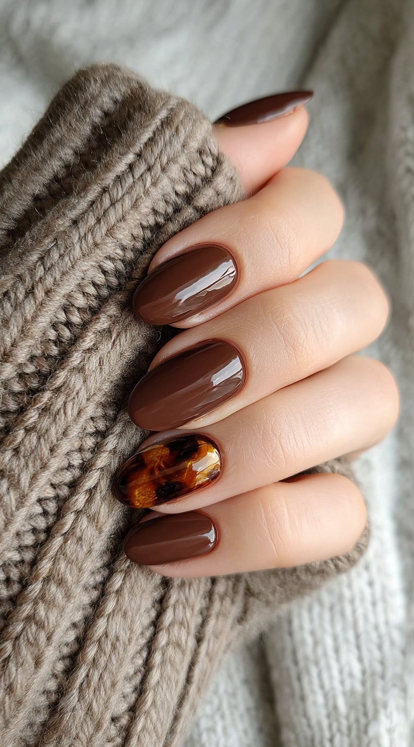 Almond-shaped nails painted in glossy chocolate brown with a tortoiseshell accent nail on the ring finger.
