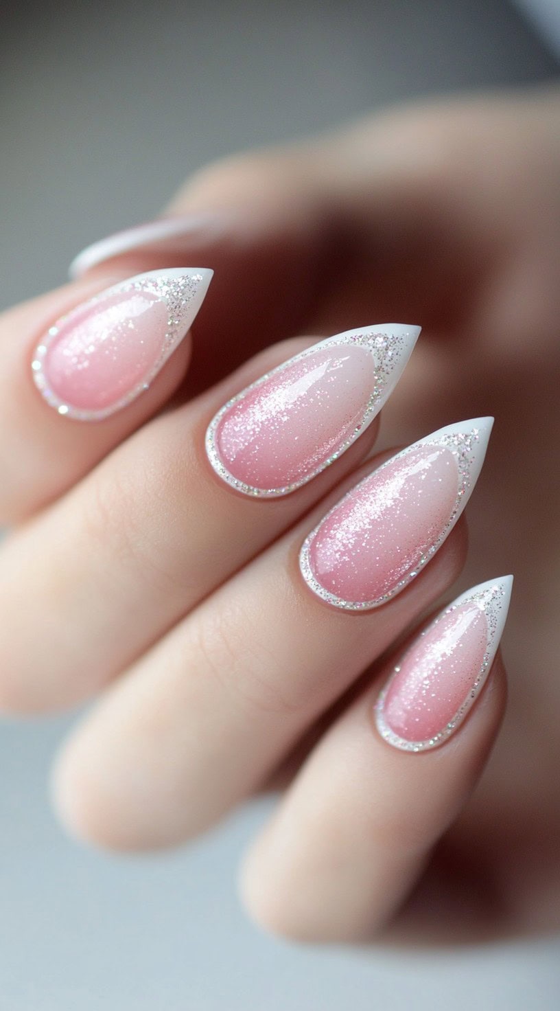A hand with almond-shaped nails featuring a soft pink ombré base, white French tips, and a sparkling glitter outline around the nail edges.