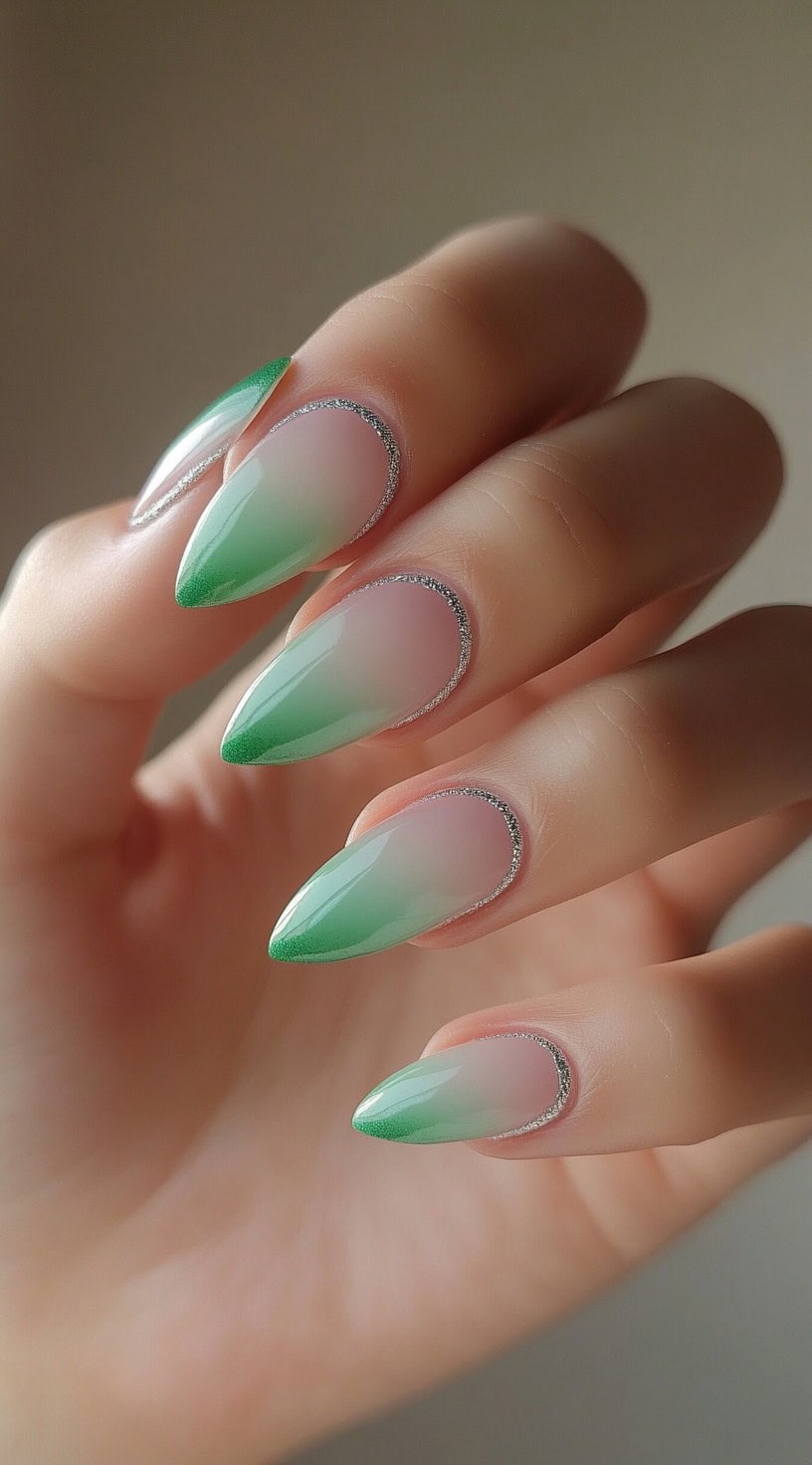 Almond-shaped nails with a soft green ombre tip and silver glitter tracing the cuticle area like a reverse French design.