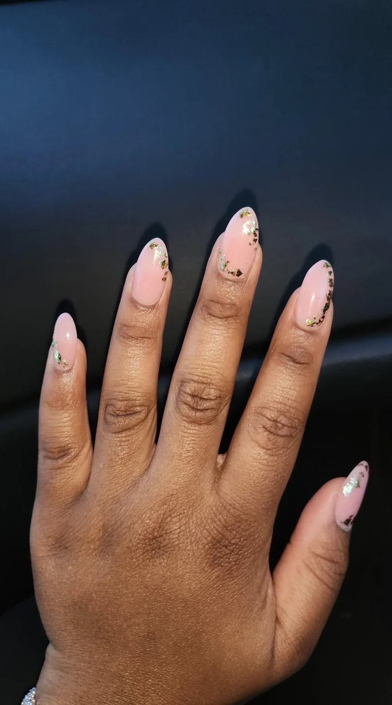 Hand with short fingers featuring almond-shaped nails painted in a glossy nude pink with shimmering gold foil accents.
