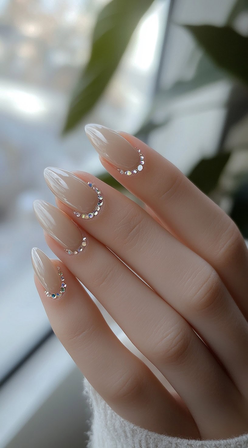 A close-up of almond-shaped nails painted in a soft nude shade, adorned with sparkling crystals elegantly placed along the cuticle.