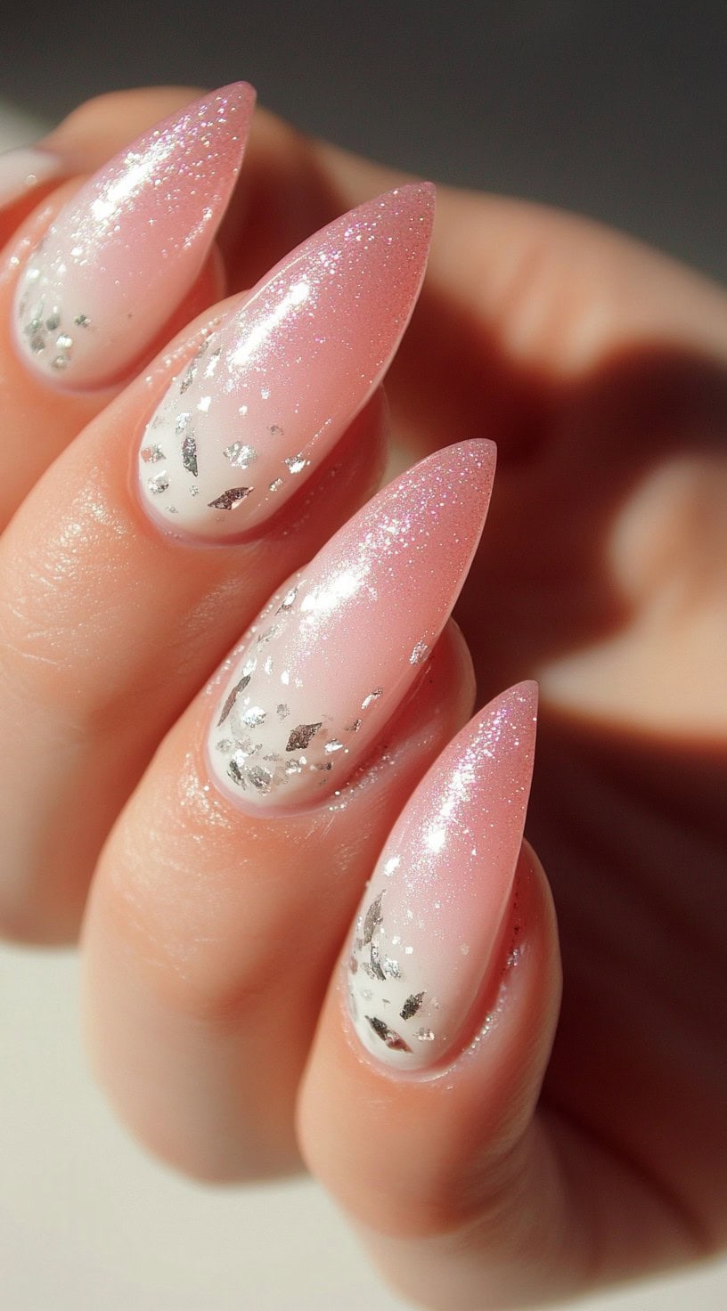 A hand with long almond-shaped nails in a pink and white ombré design with silver foil flakes scattered across the tips.