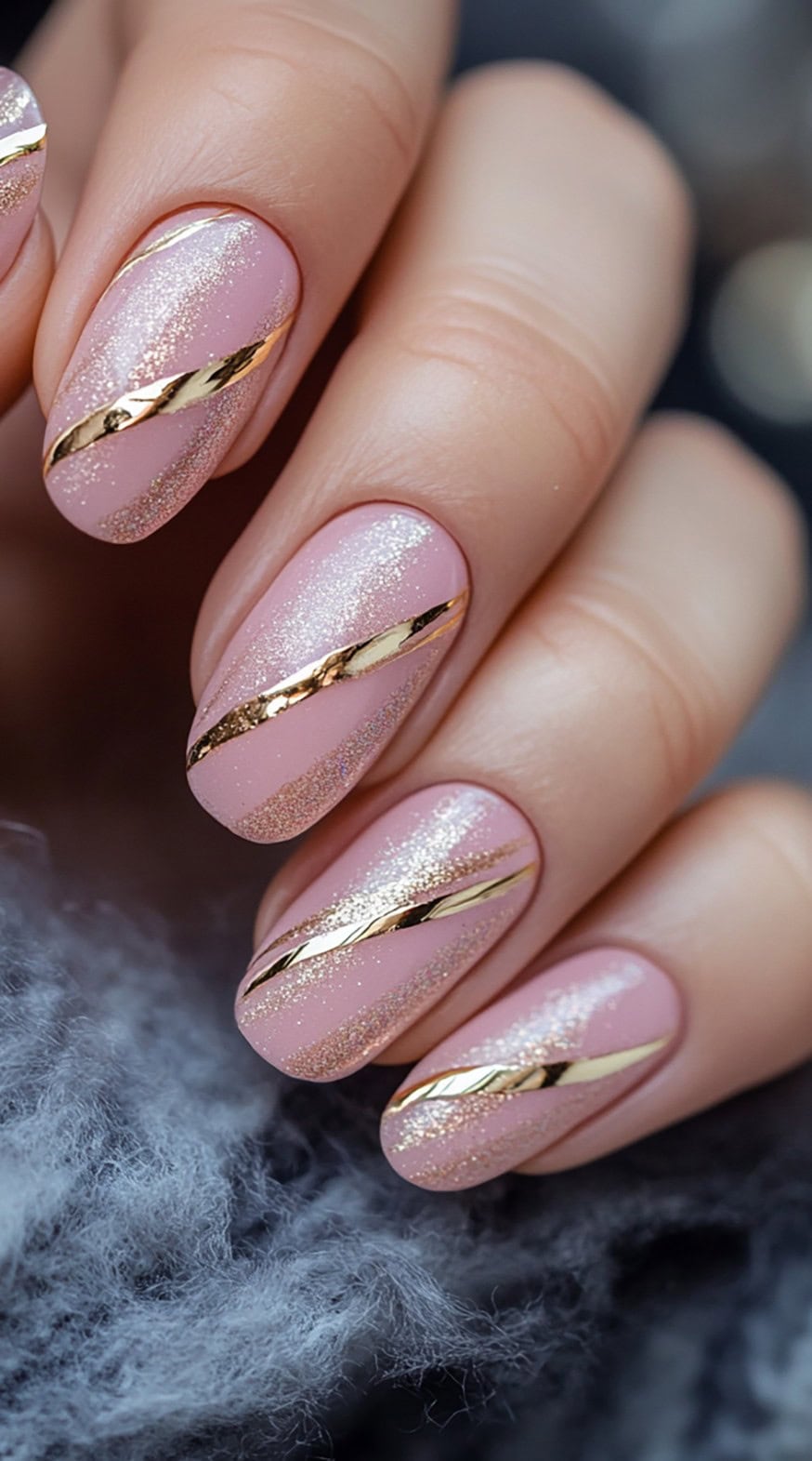 A set of almond-shaped nails with a soft pink base, dusted with glitter and adorned with diagonal gold foil stripes.