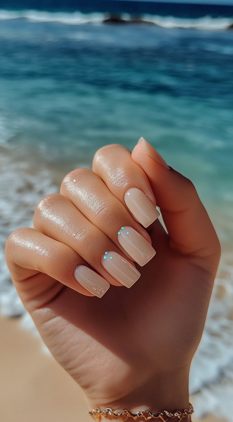 Hand with square-shaped nude nails featuring tiny aqua dots near the cuticles.