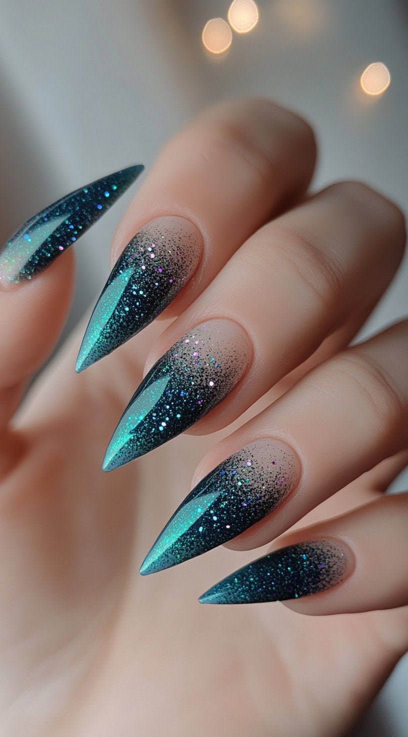 A hand with sharp stiletto nails fading from clear base to teal glitter with specks of multicolor shimmer.
