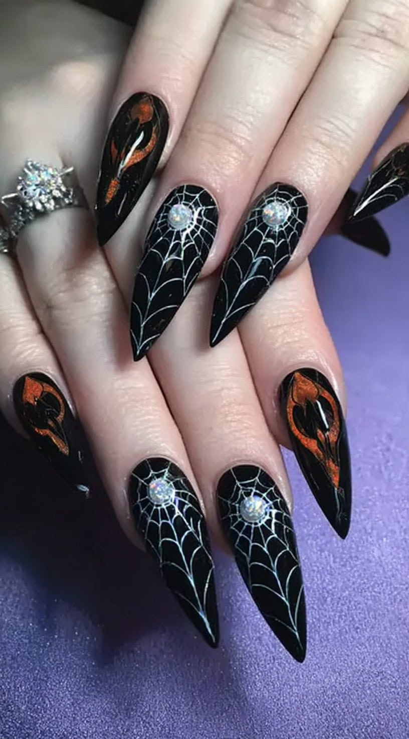 A hand with sharp stiletto nails painted black, featuring white spider webs and rhinestones at the web centers, along with glowing flame accents.