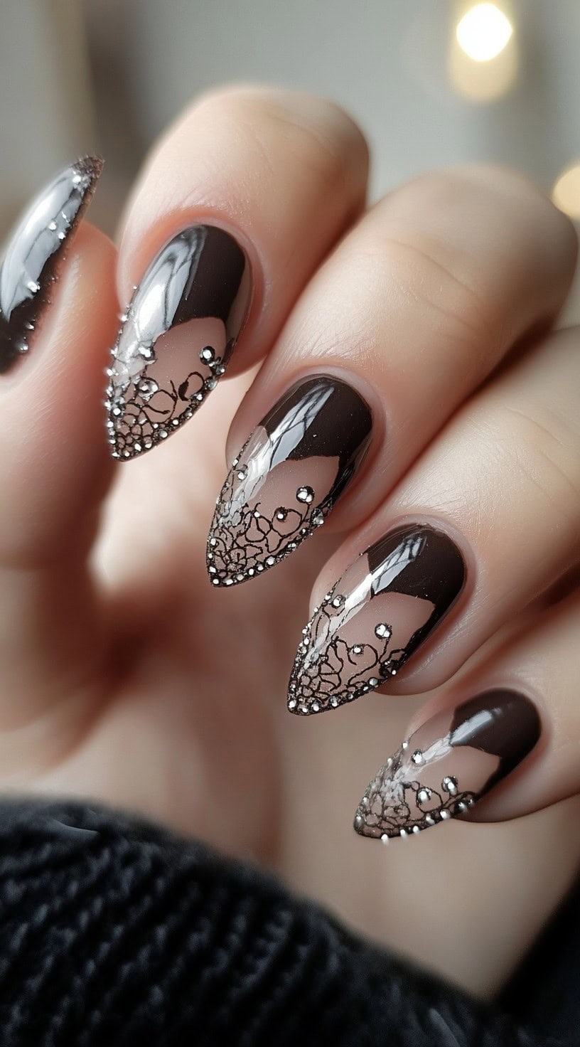 Stiletto-shaped nails with sheer nude bases, glossy deep brown tips, and delicate black lace designs with tiny rhinestones.