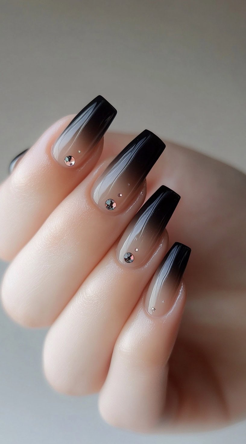 A set of long, square-shaped nails with a gradient ombre effect transitioning from sheer nude to deep black, embellished with small rhinestones for added sparkle.