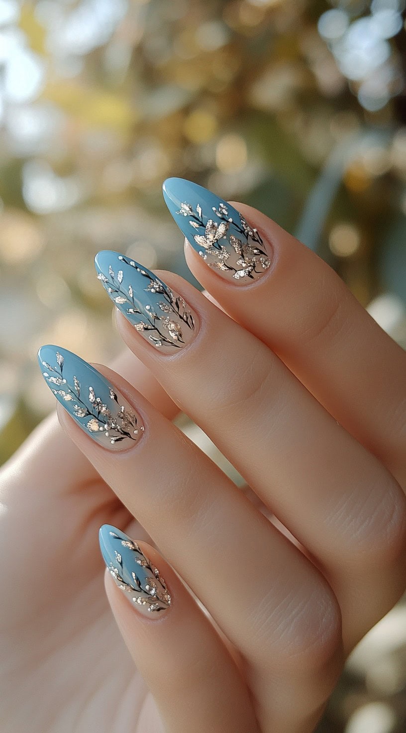 A close-up of almond-shaped nails with a blue and nude gradient base, adorned with delicate silver floral accents.