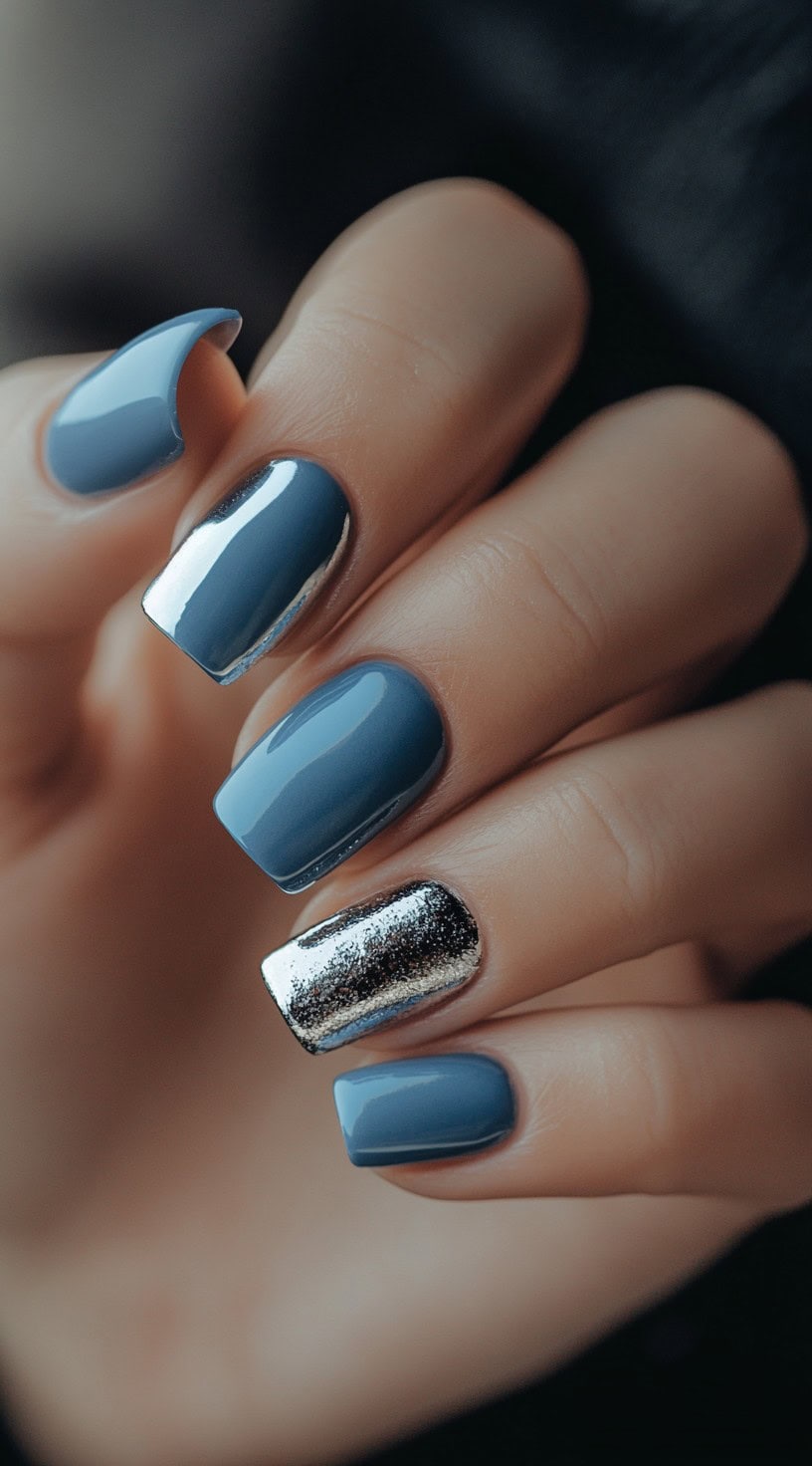 A hand with square-shaped nails painted in a cool blue chrome, featuring one accent nail with silver foil and glitter.
