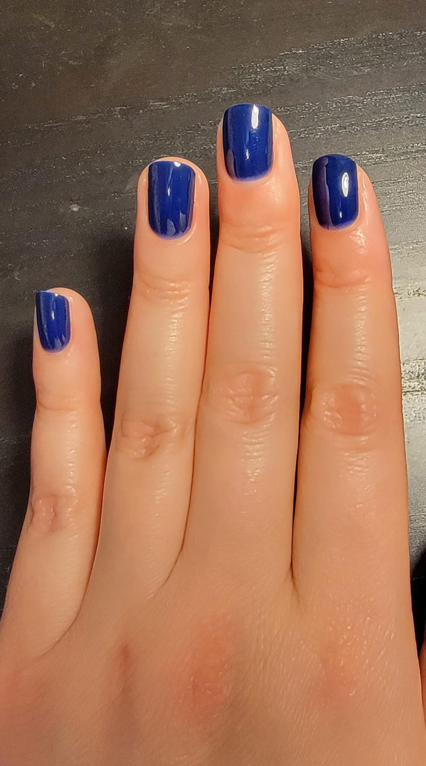 Hand with short square nails painted in a bold, glossy blue gel polish, resting on a black surface.