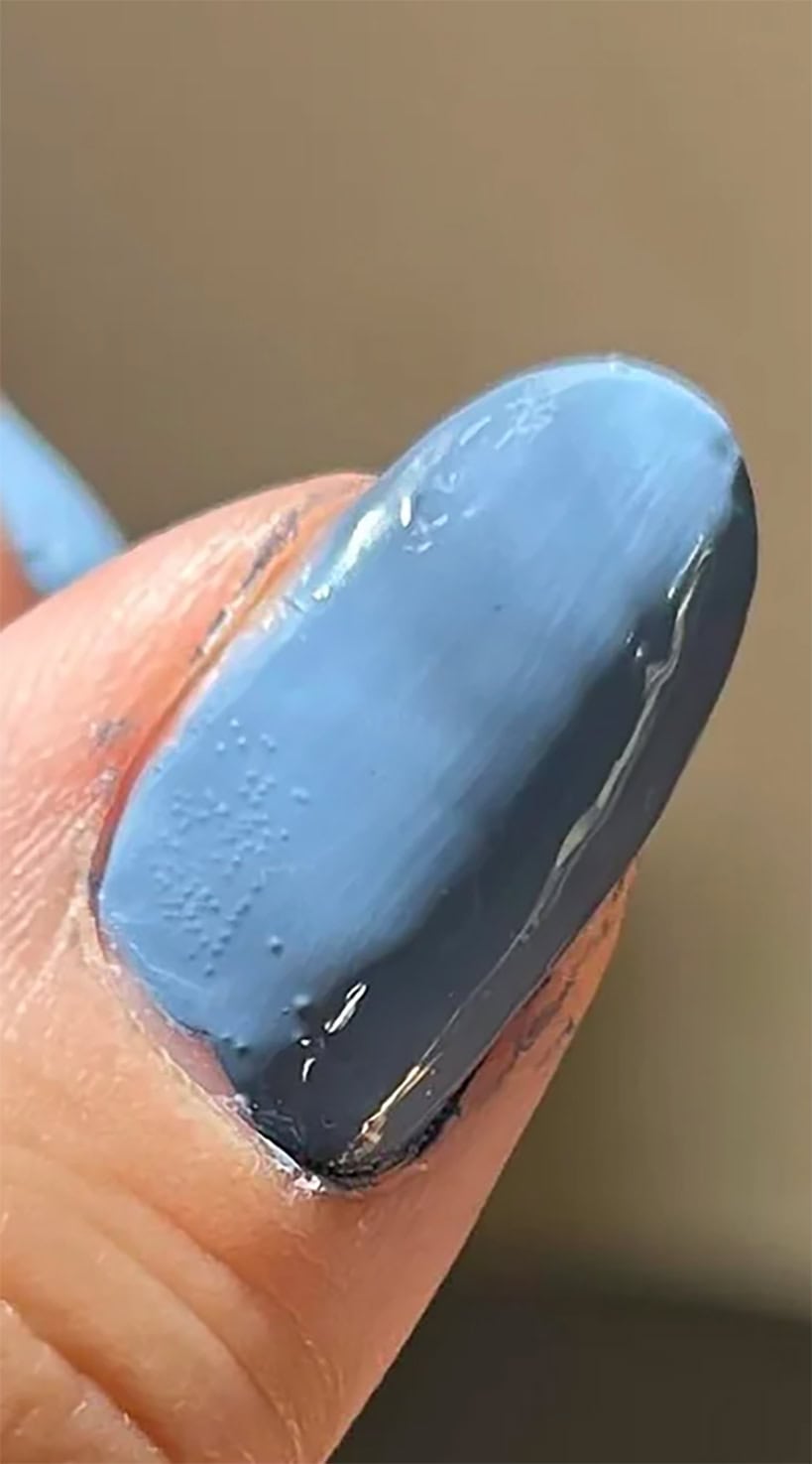 Blue-painted thumbnail showing tiny air bubbles and rough spots under the polish, especially near the cuticle area.