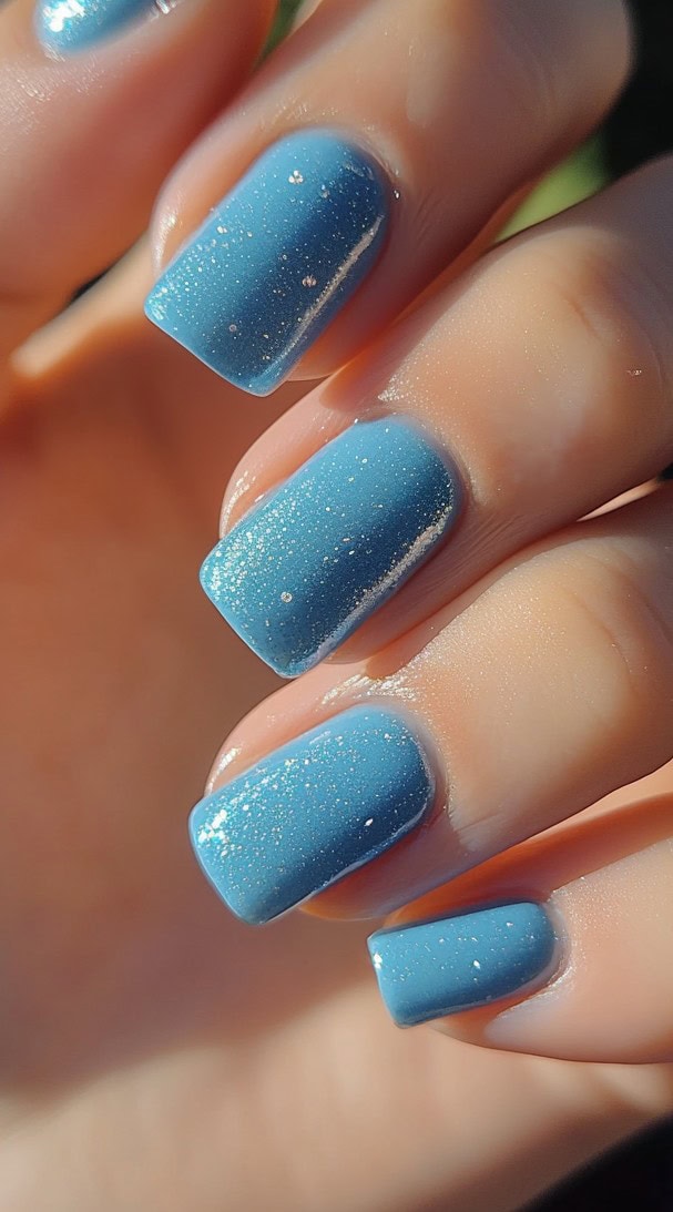 A hand with medium-length square nails painted in a vibrant blue polish with a fine layer of iridescent glitter.