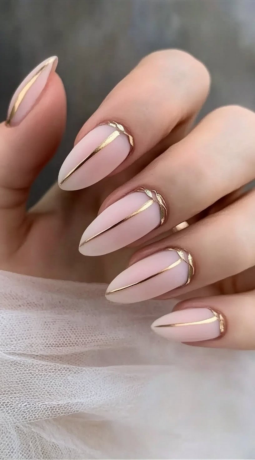 A close-up of almond-shaped nails with a soft blush pink matte base, featuring sleek gold lines for a minimalist yet elegant design.