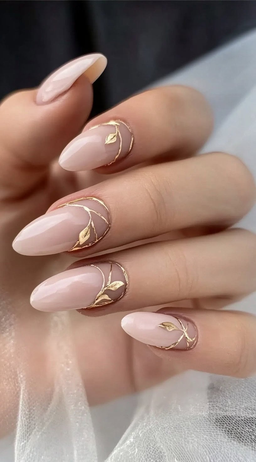 A close-up of a hand with almond-shaped nails in a soft blush nude tone, decorated with fine gold leaf detailing near the cuticles.