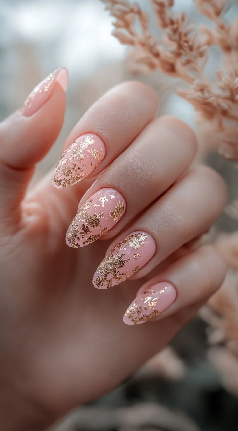 A hand with almond-shaped nails painted in a soft blush pink shade, adorned with scattered gold leaf accents.