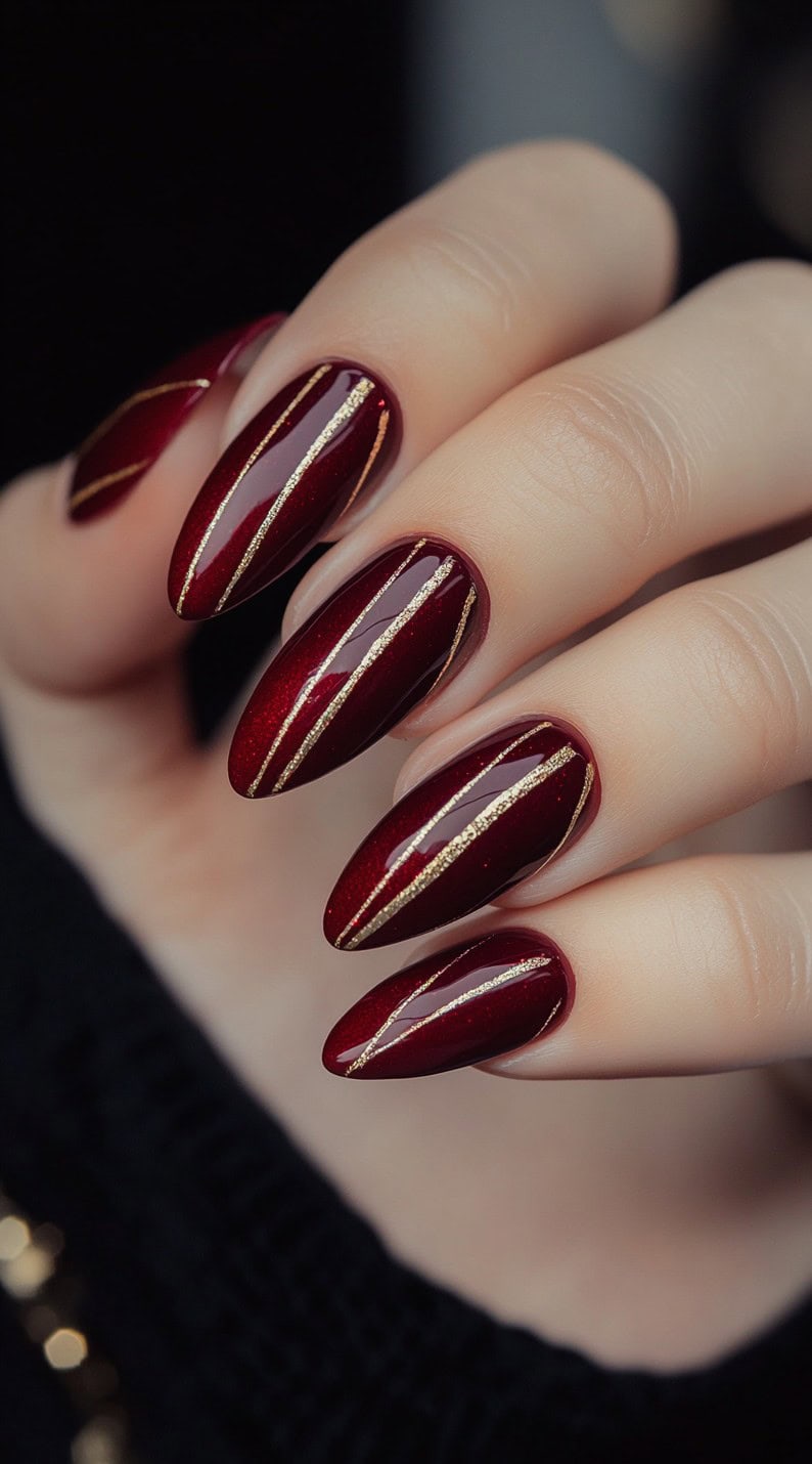 A set of almond-shaped burgundy nails with thin, vertical gold striping, creating a sleek and sophisticated bridal nail look.