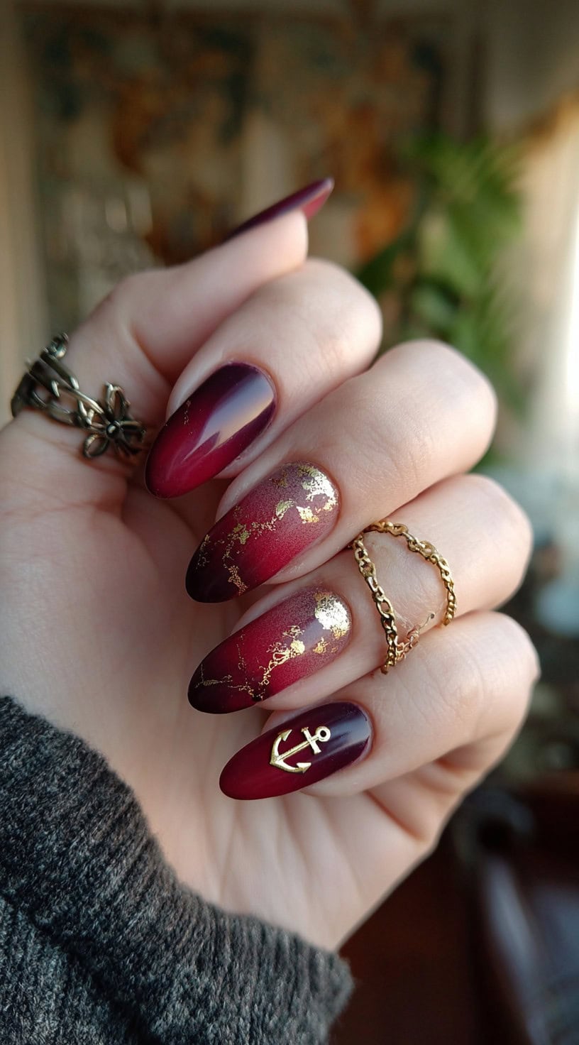 Elegant almond-shaped nails in a burgundy ombré are enhanced with gold foil accents and a single gold anchor charm.