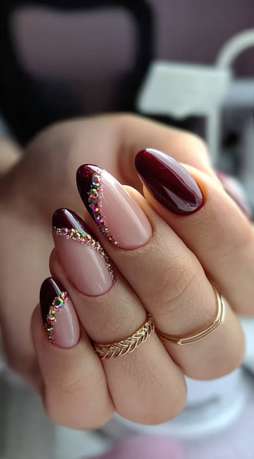 A set of almond-shaped nails featuring nude bases, burgundy French tips, and rhinestone curves along the tip line.