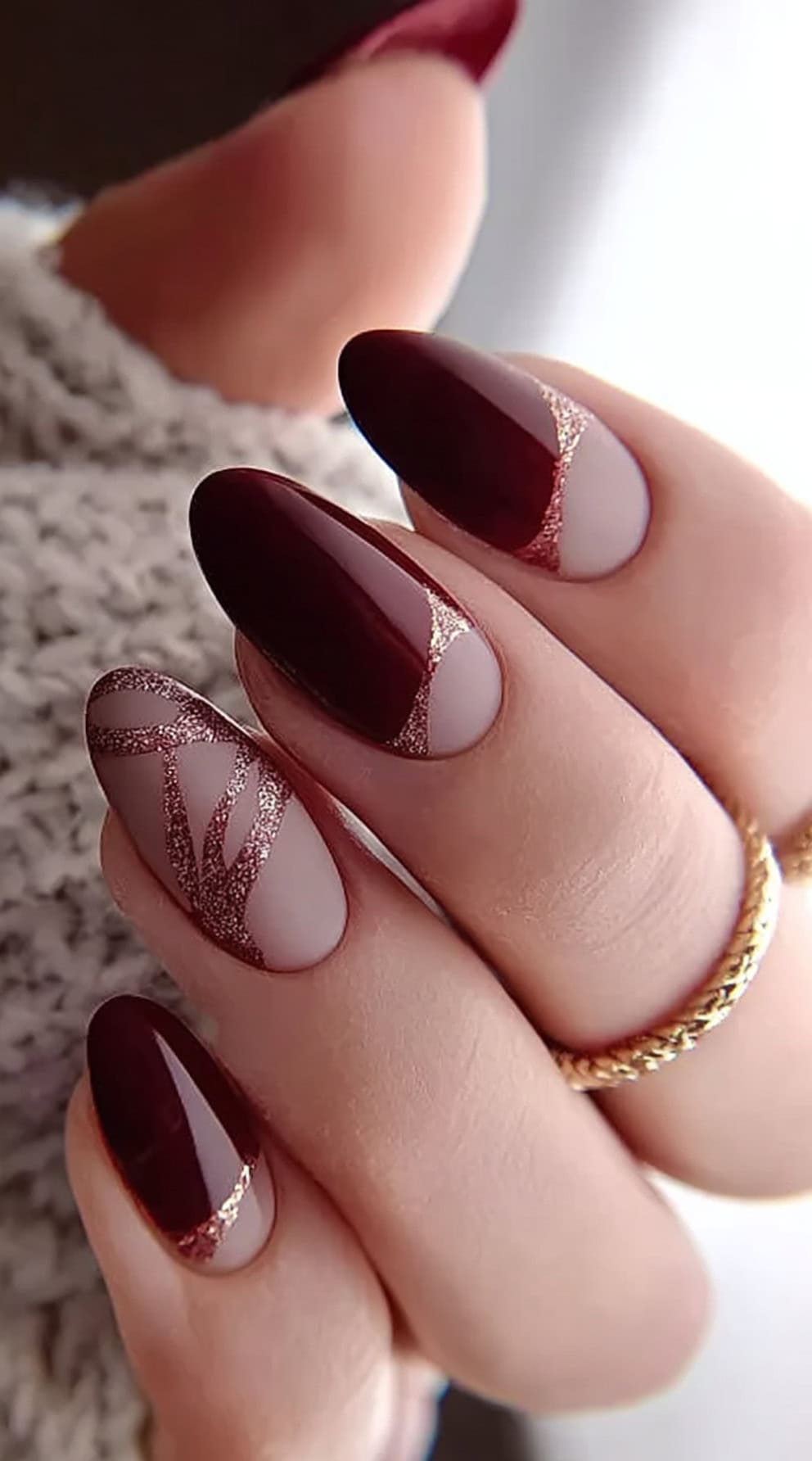 A hand with almond nails featuring burgundy French tips outlined in rose gold glitter, paired with linework on a nude base.