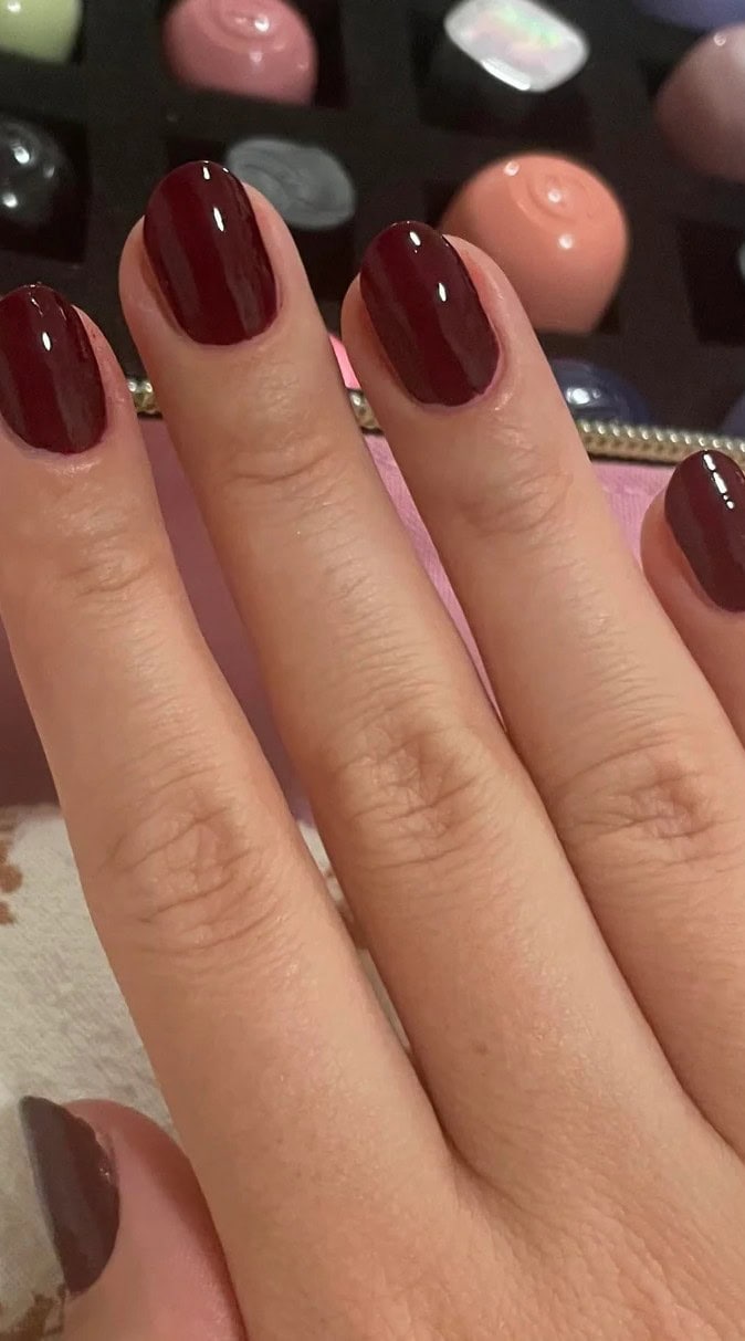 Dark burgundy gel-polished nails with a reflective shine, displayed against a background of nail polish bottles.