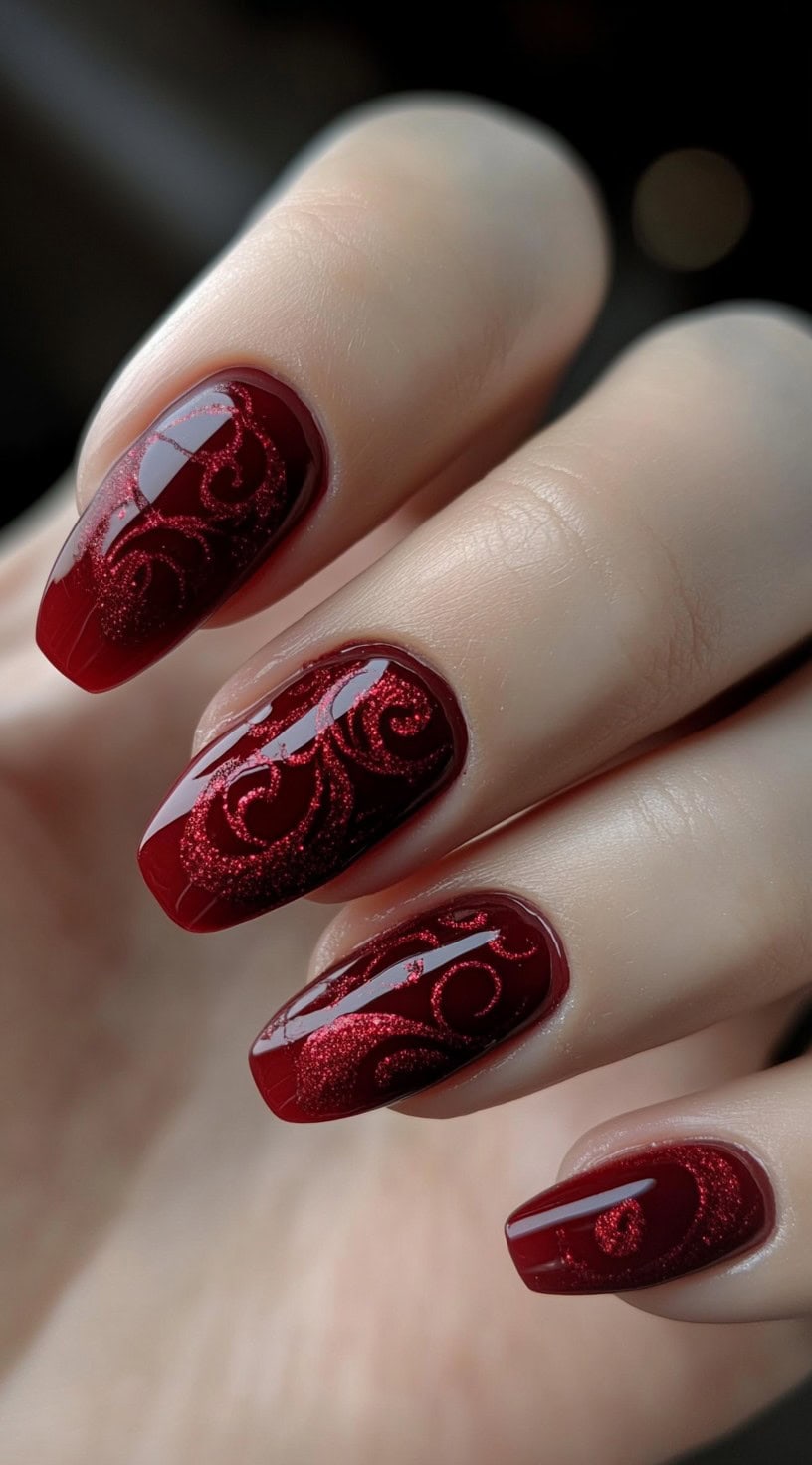 Medium-length oval nails painted in glossy burgundy with fine red glitter scrollwork across the surface.