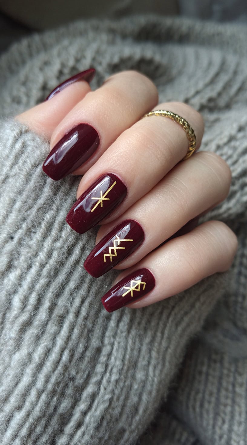A hand with long burgundy coffin-shaped nails, each featuring minimalist gold rune details.