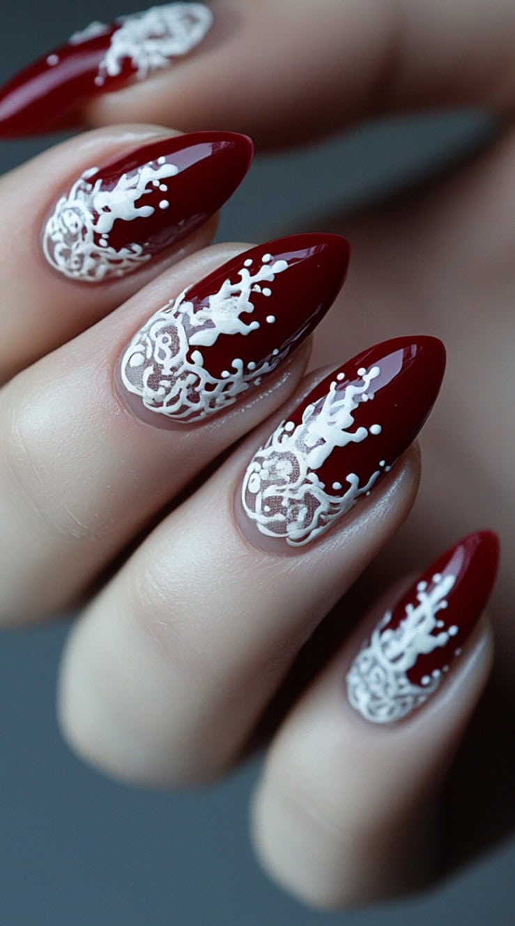 Elegant burgundy nails featuring intricate white lace patterns delicately painted onto an almond shape.