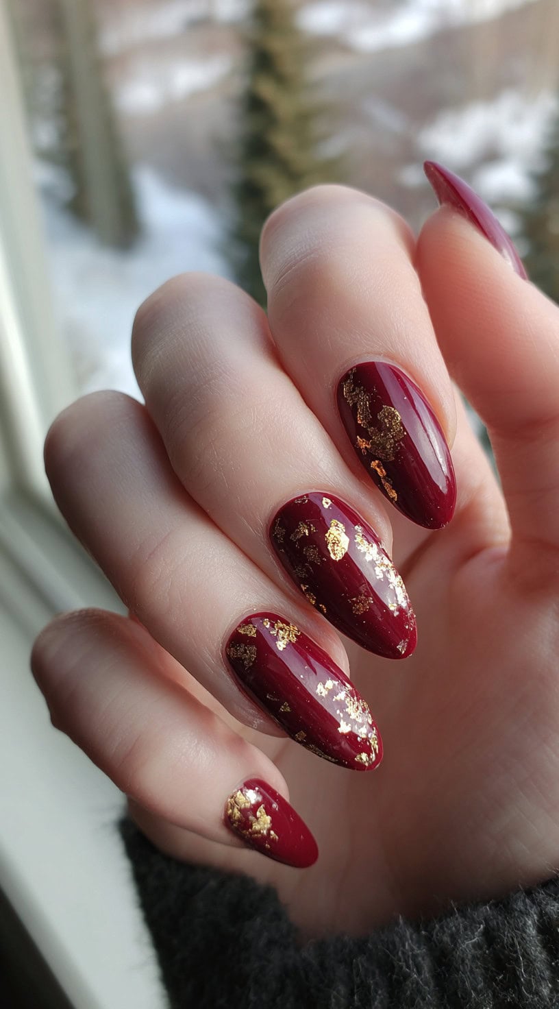 A close-up of almond-shaped nails painted in glossy burgundy with clustered metallic gold foil flakes on top.
