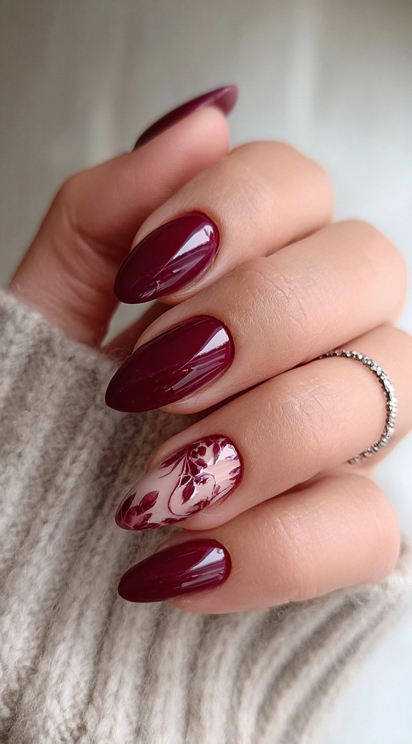 A hand with deep burgundy nails and one accent nail decorated with a burgundy floral design over a milky nude base.