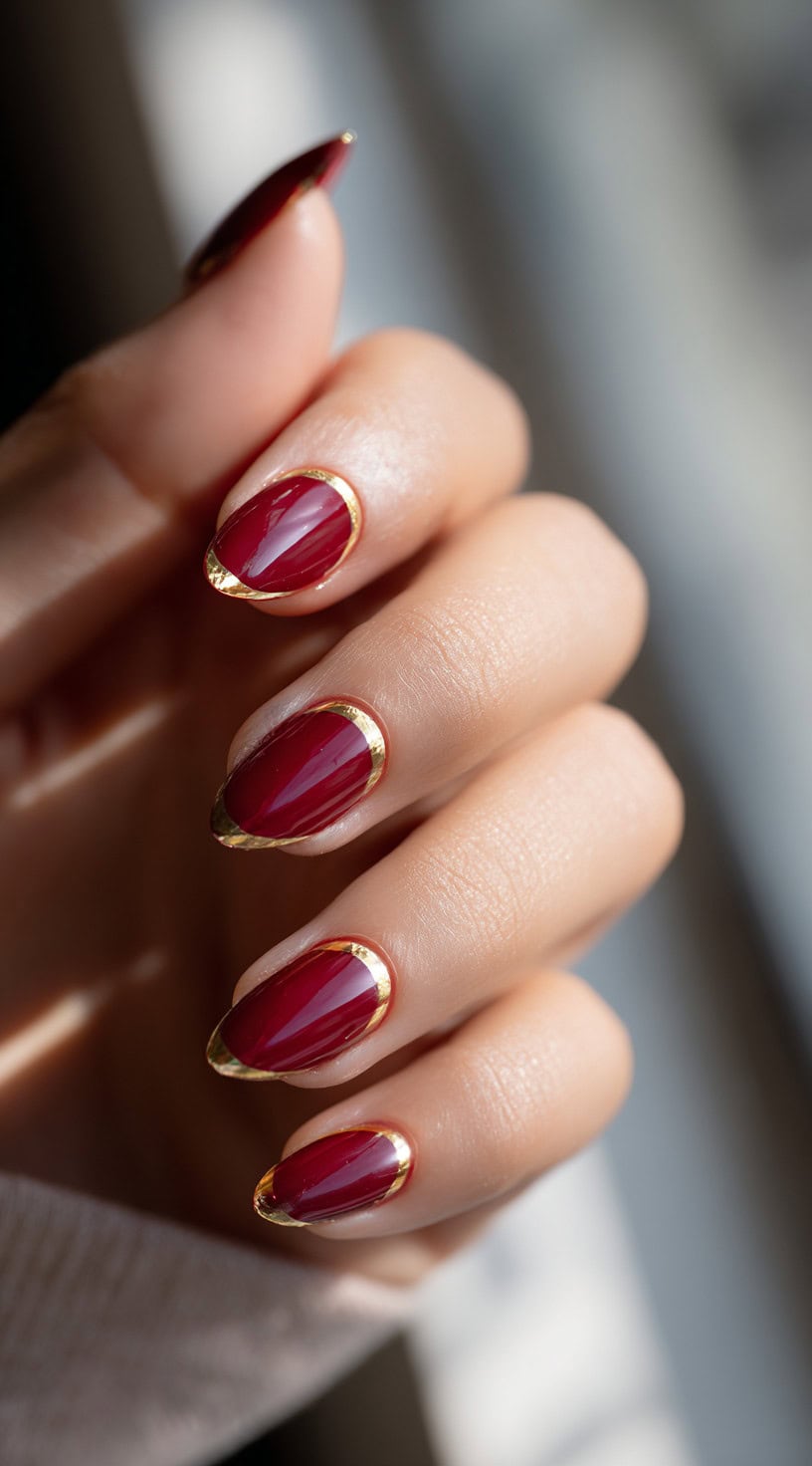 A close-up of almond-shaped nails in rich burgundy polish bordered with gold foil along the edges and cuticles.