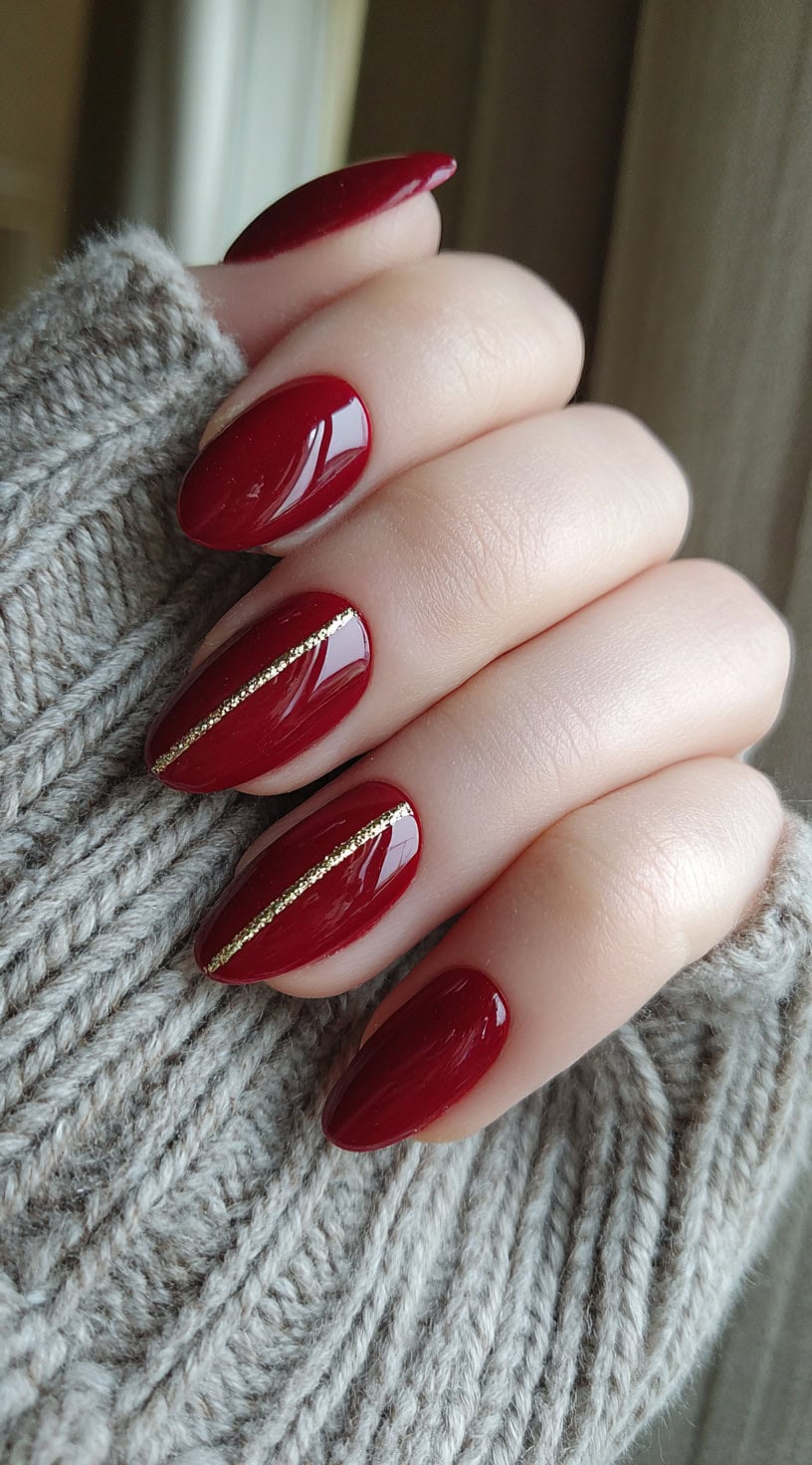 A hand with burgundy almond nails, each nail featuring a clean vertical gold stripe down the center.