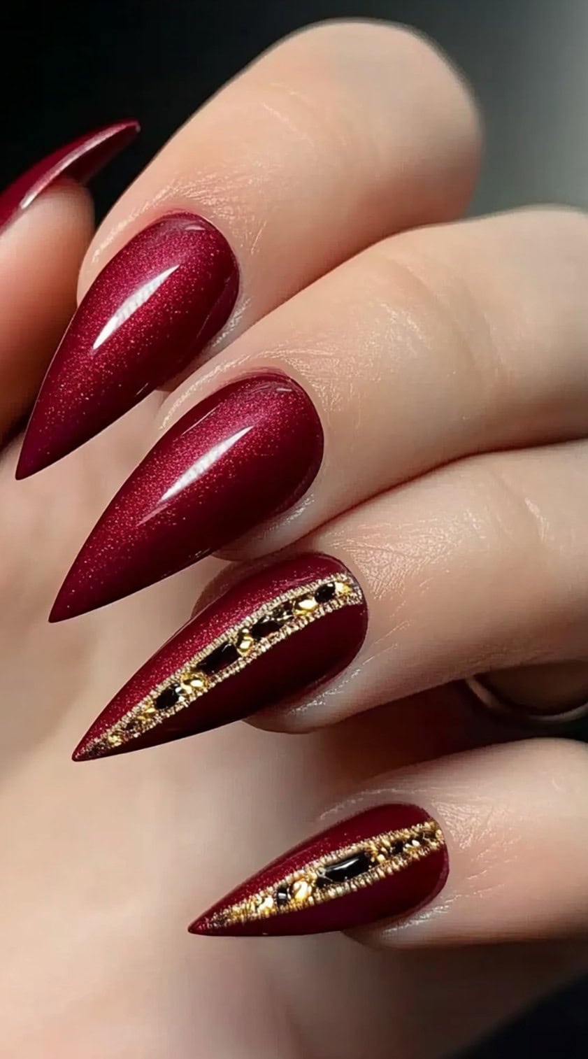 Deep burgundy stiletto nails with a shimmer finish, featuring gold-lined slits filled with black and gold gemstones.