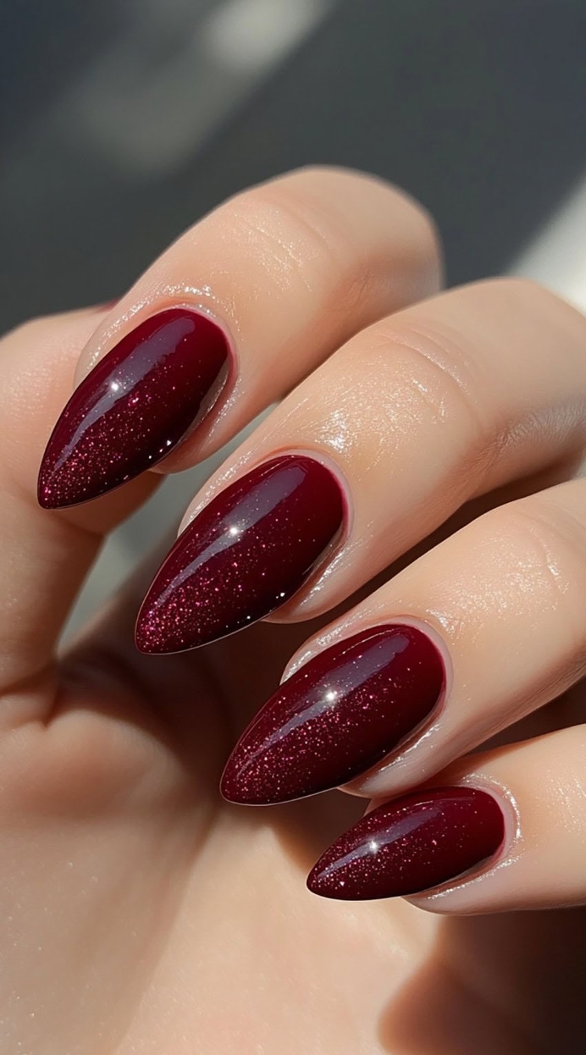 Glossy almond-shaped nails in a deep burgundy tone with a subtle sparkle effect.