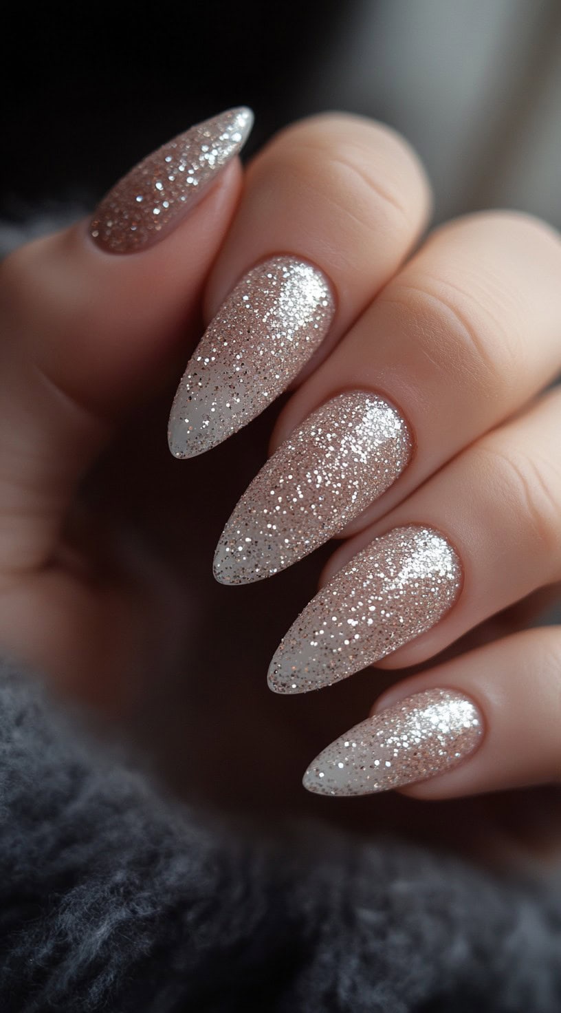 A hand with almond-shaped nails featuring a nude-to-champagne glitter ombre effect, sparkling under soft lighting.