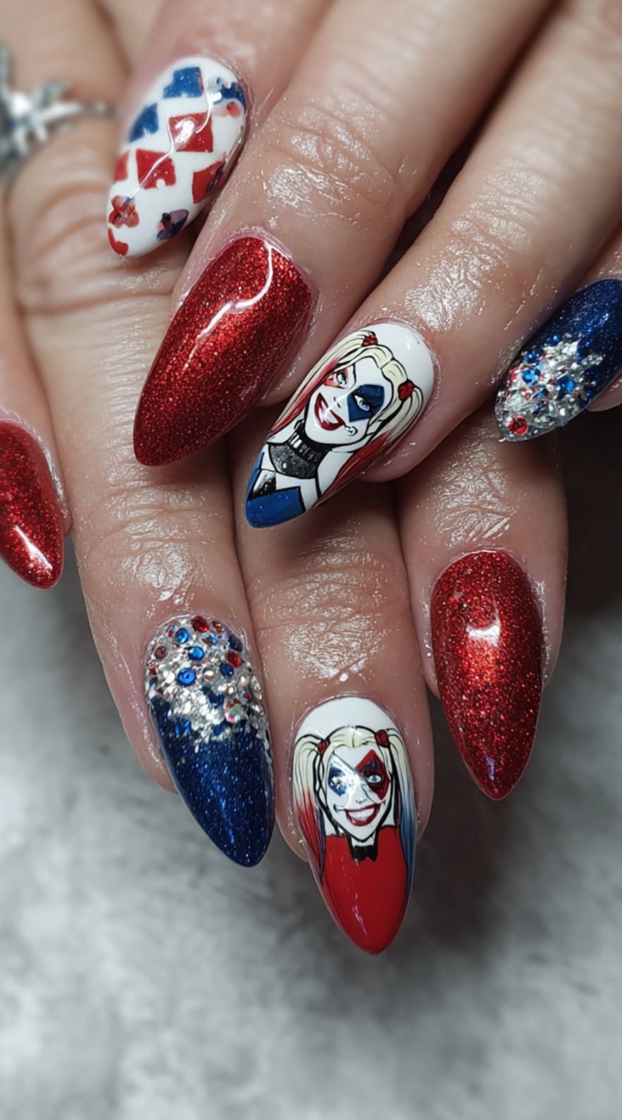 Almond nails with red and blue glitter, rhinestones, and painted portraits of Harley Quinn's comic-style face.