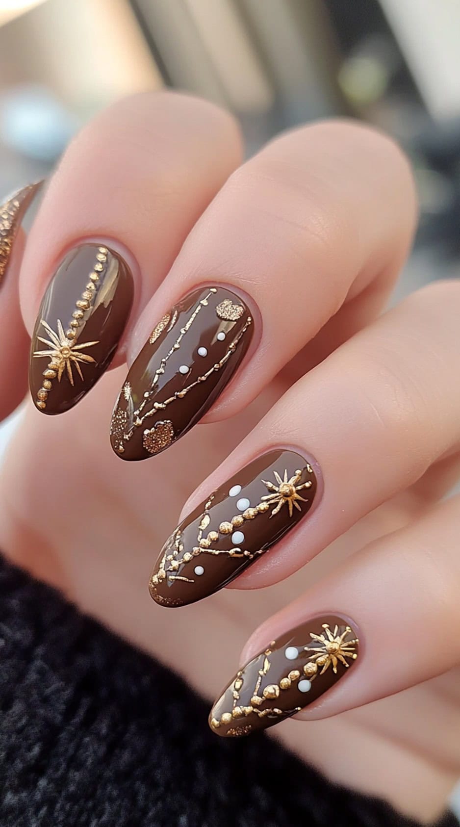 Almond-shaped brown nails featuring gold celestial designs, including stars, dots, and delicate swirling patterns.