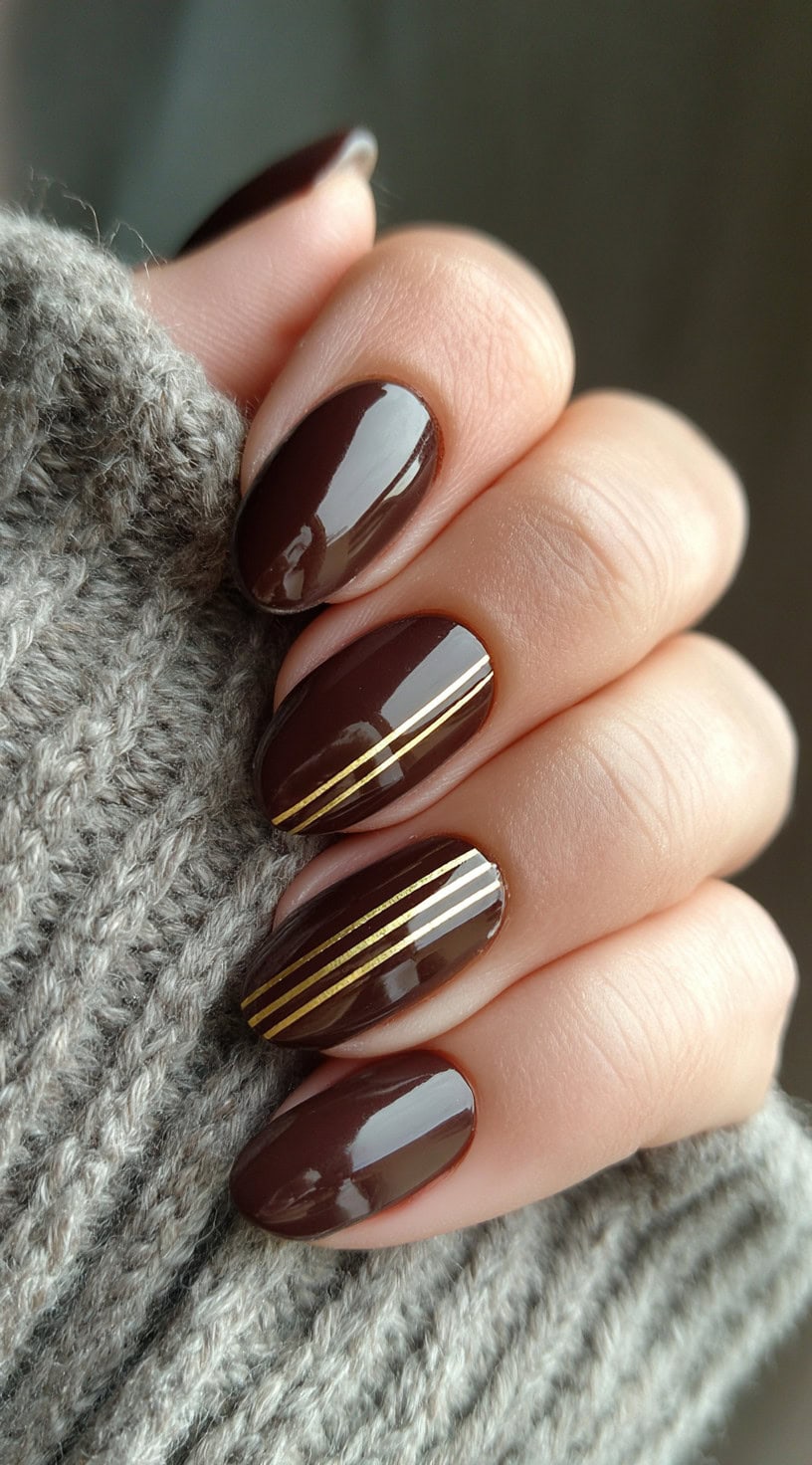 Glossy chocolate brown oval nails with two thin gold metallic stripes on accent fingers.