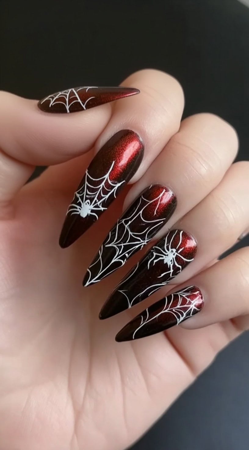A hand with stiletto nails in a deep red chrome finish, each decorated with white spiderwebs and tiny white spiders.