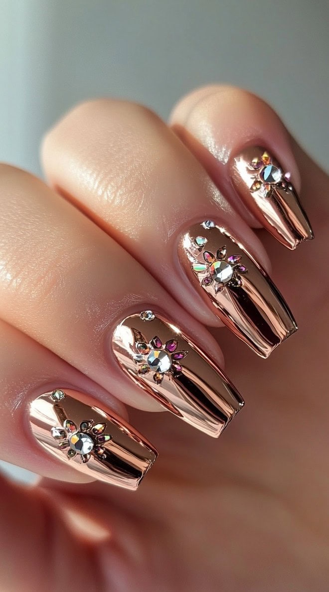 A hand with long, almond-shaped nails coated in reflective rose gold chrome polish, adorned with sparkling crystal embellishments.