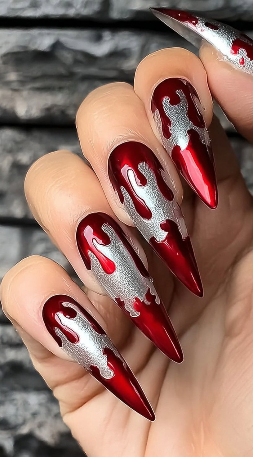 A hand with long red chrome stiletto nails featuring silver metallic blood drip accents over a deep red base.