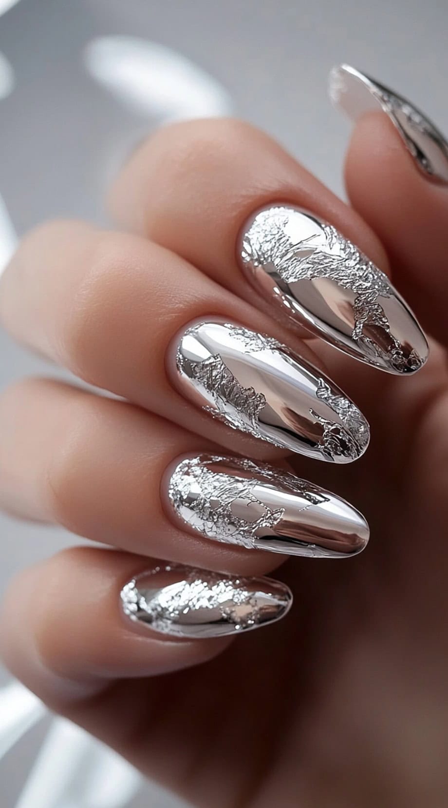 A close-up of almond-shaped nails with a reflective chrome silver finish, accentuated with delicate silver foil details.