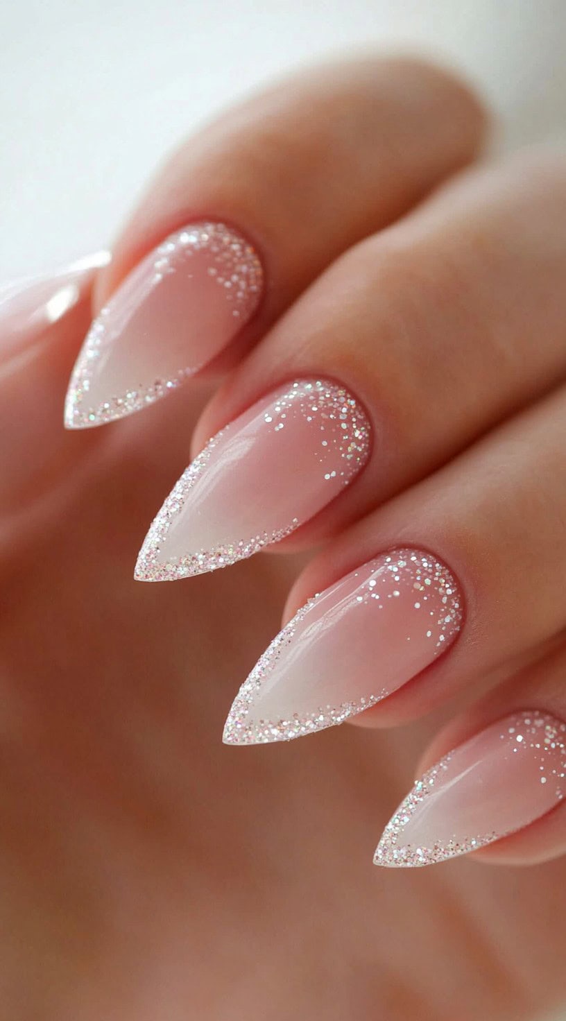 A hand with almond-shaped nails in a pink ombré style, outlined with a fine glitter border along the edges and tips.