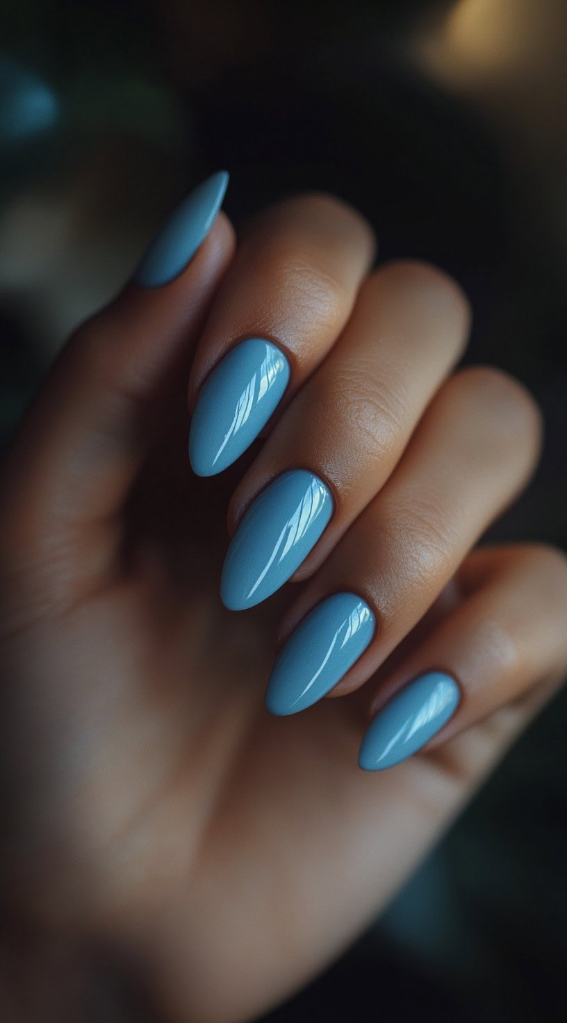 A close-up of a hand with glossy, almond-shaped nails painted in a muted dusty blue shade against a dark, moody background.