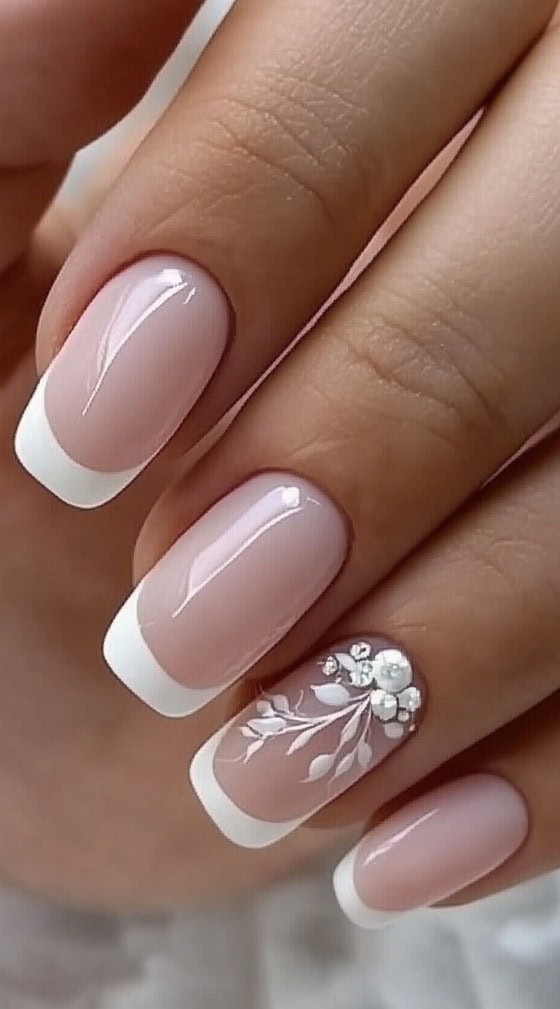 A close-up of a hand with classic French manicure nails, featuring one accent nail with delicate white floral patterns and pearl embellishments.