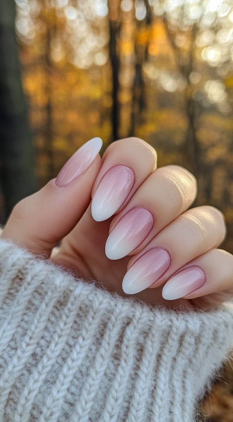 A hand with almond-shaped nails featuring a seamless pink-to-white ombre design, set against an autumnal background with golden leaves.